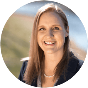 Sarah-Grace Parr smiling in front of a blurred background of grass and a pond. She is wearing a pearl necklace and a blazer.