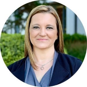 Liz Lee wears a black coat and lightweight grey top with a shawl collar. She has blonde hair and stands in a natural setting