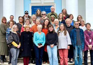 A divers group of about 25 Death Doulas in casual clothing stand on steps in front of a blue door and blue walls.